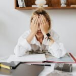 A woman sits at a home office desk overwhelmed, with her face in her hands, depicting stress and frustration.