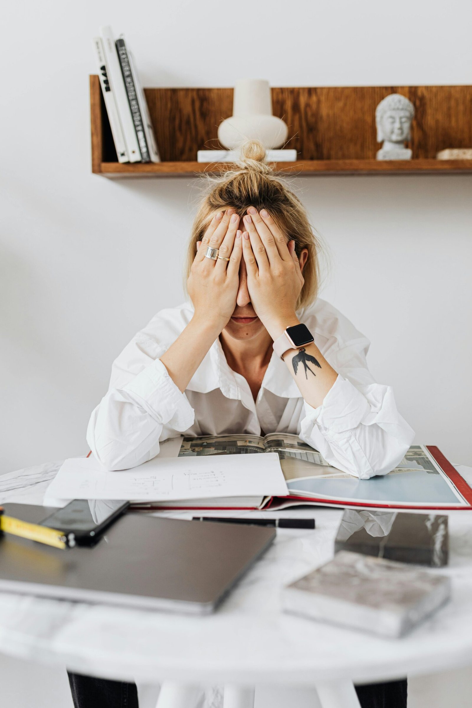 A woman sits at a home office desk overwhelmed, with her face in her hands, depicting stress and frustration.