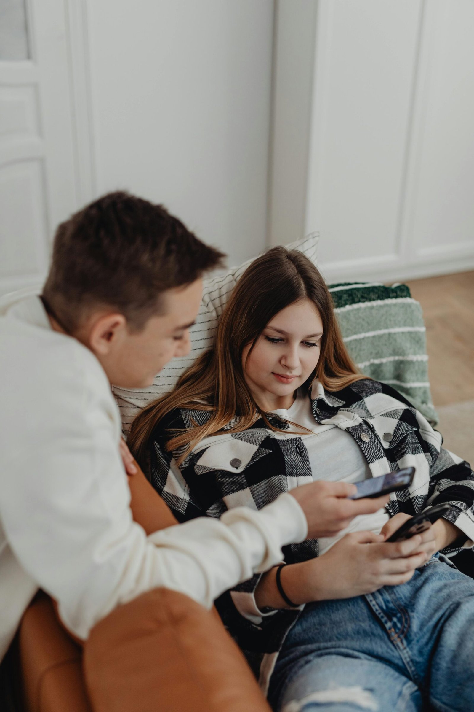 Two teenagers lounging indoors, engaging with smartphones and sharing content.