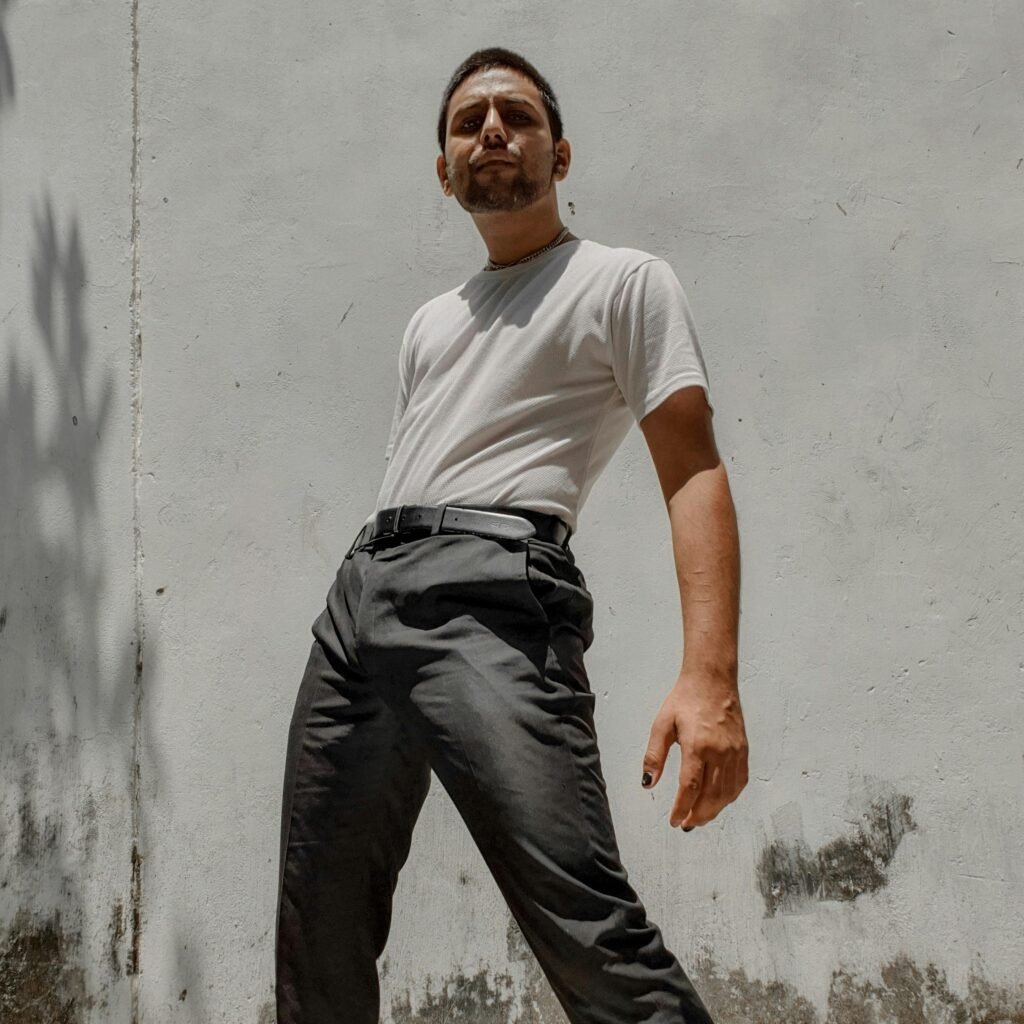 A stylish young man striking a confident pose against a rustic white wall outdoors.